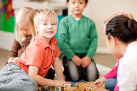 A young man looks up smiling tentatively among a group of children playing with blocks on the floor