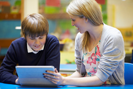 Teacher sitting beside student at table demonstrating how to use app on digital tablet